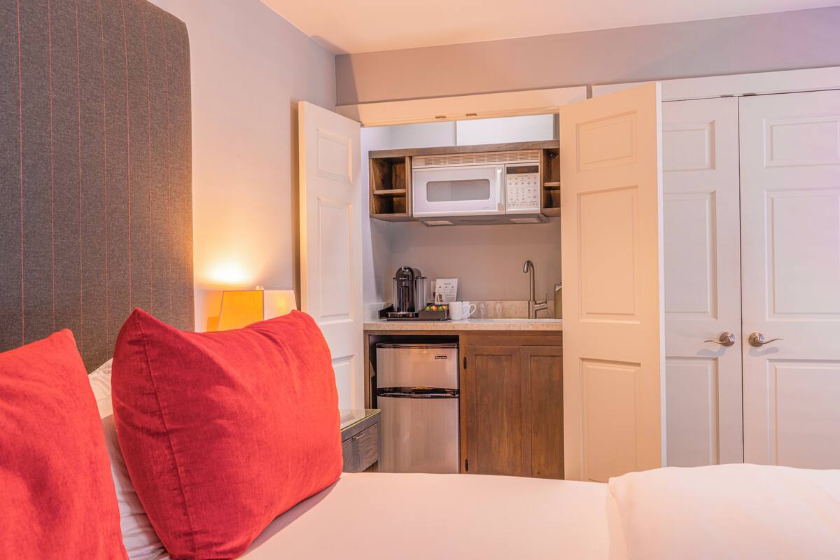 A compact hotel room with a kitchenette, a bed with red pillows, and a white wardrobe.