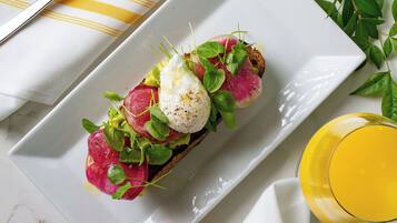 Une assiette rectangulaire blanche sur laquelle on a disposé des œufs sur des toasts, des légumes verts et des radis, accompagnée d’une fourchette et d’un couteau posés sur une serviette rayée jaune et blanche.