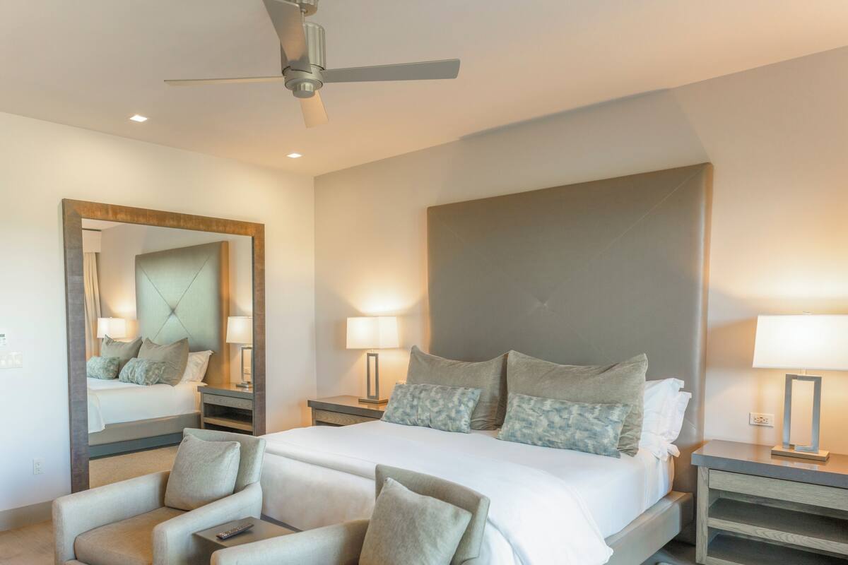 A modern hotel room with a large bed, two bedside tables with lamps, a ceiling fan, a seating area with chairs, and a large mirror.