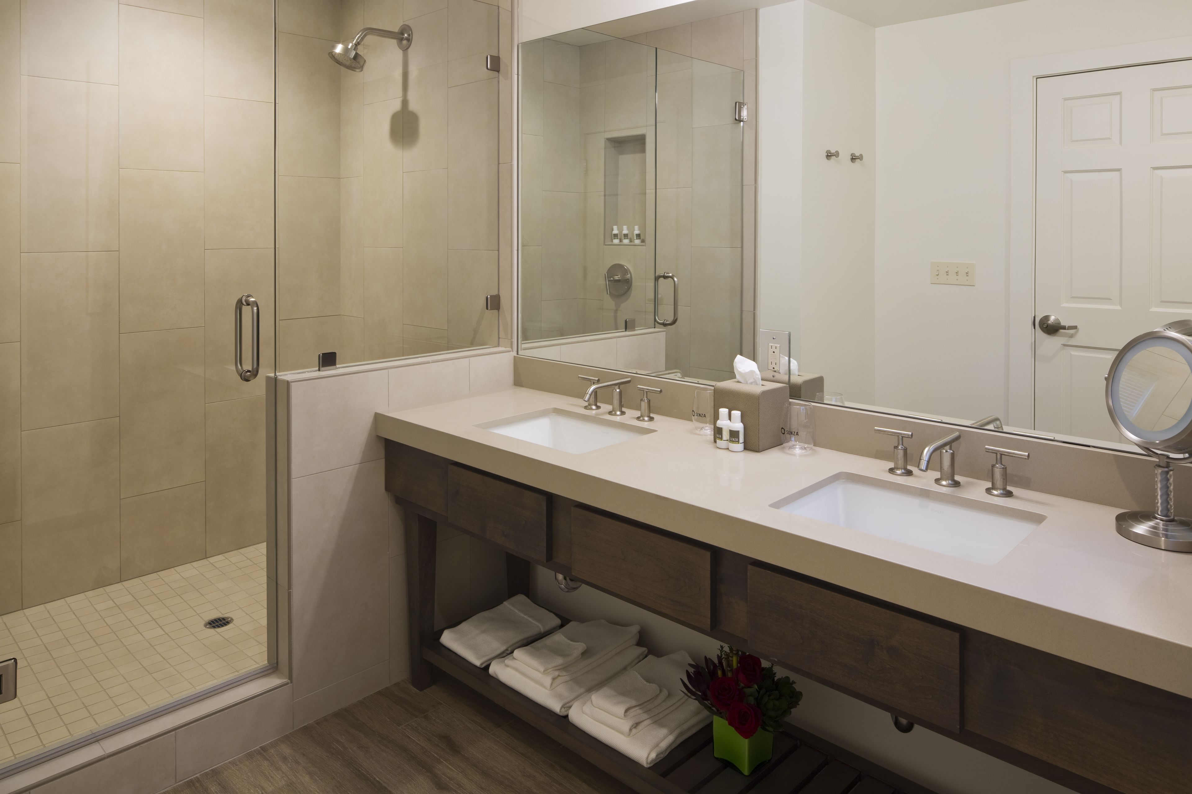 A modern bathroom with a walk-in shower, two sinks, a large mirror, and a vanity with storage drawers.