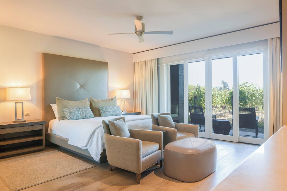 A modern hotel room with a large bed, two armchairs, a round ottoman, and a ceiling fan.