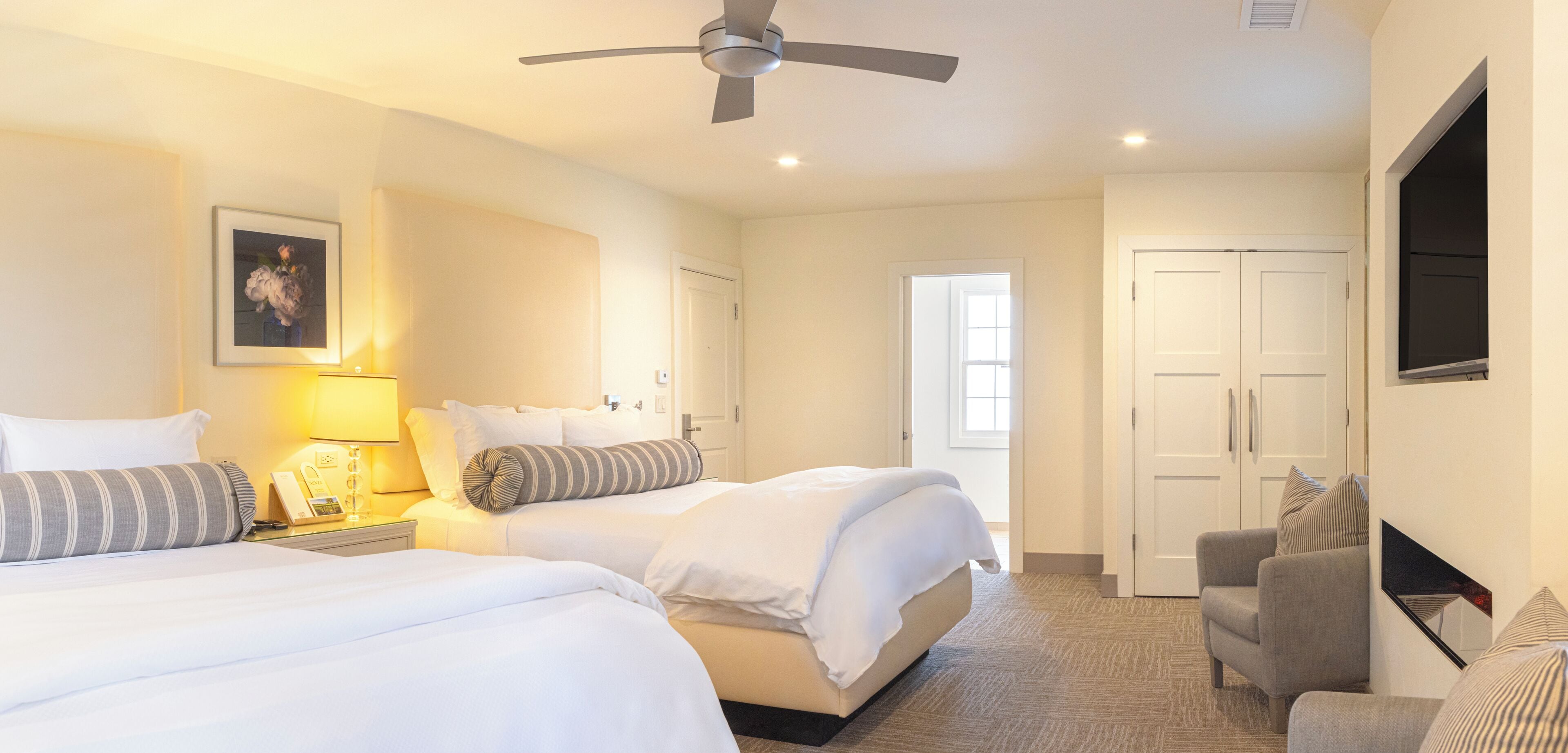 Habitación de hotel con dos camas, un escritorio con lámpara, una silla, un ventilador de techo y una puerta que da a otra habitación.