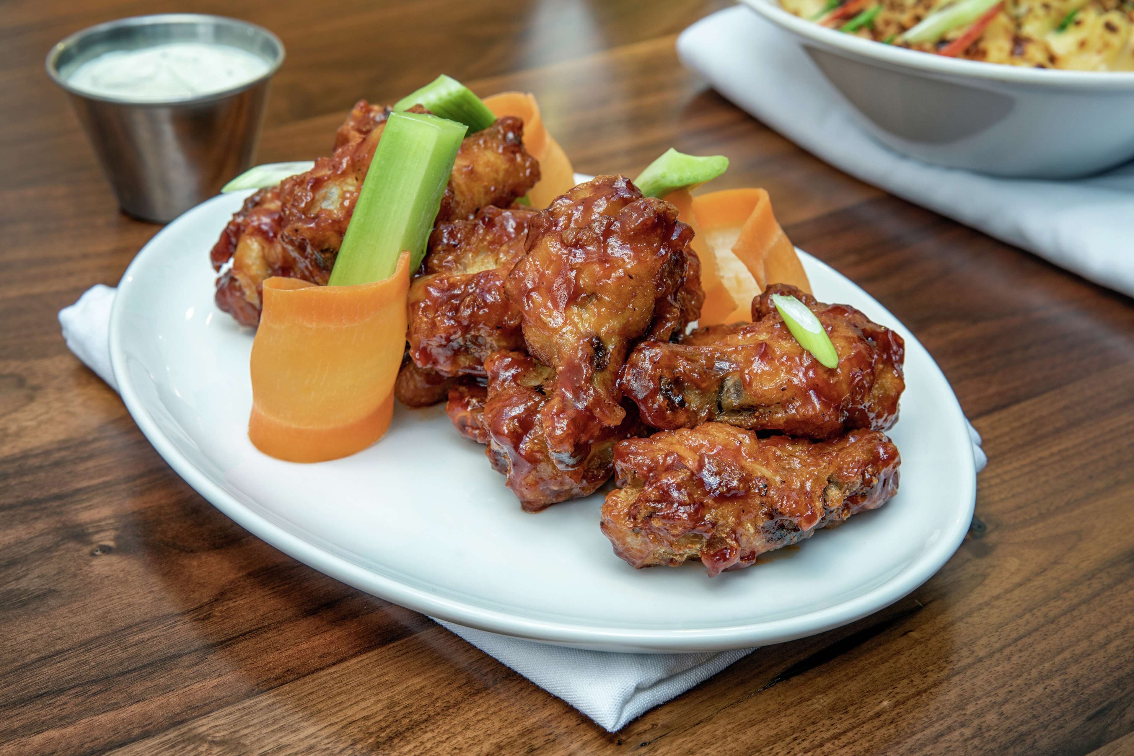 A plate of chicken wings with celery and sauce, a small metal cup, and a bowl of salad in the background.