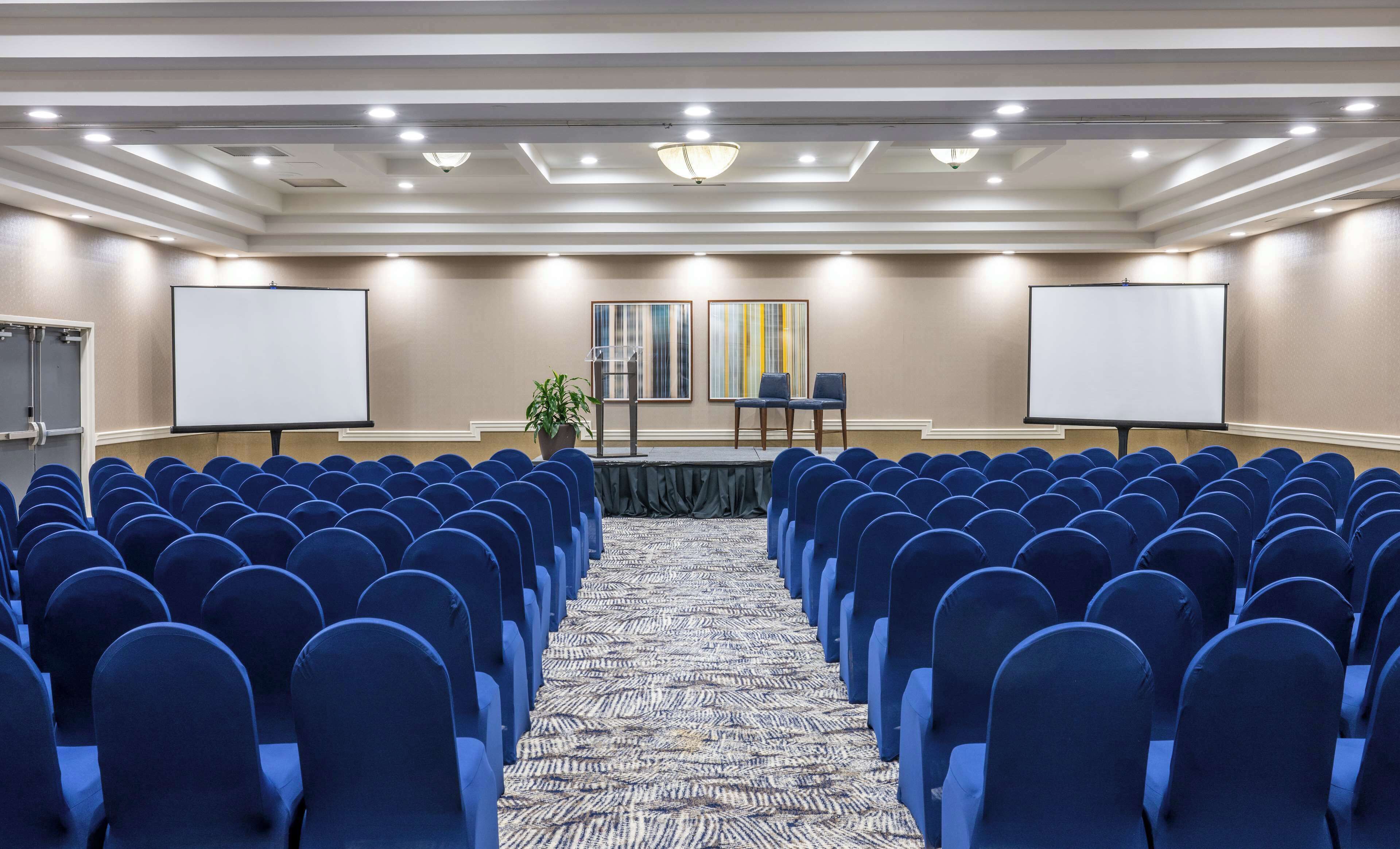 Un salón de conferencias con filas de sillas azules, dos pizarras blancas, un atril y una alfombra estampada.