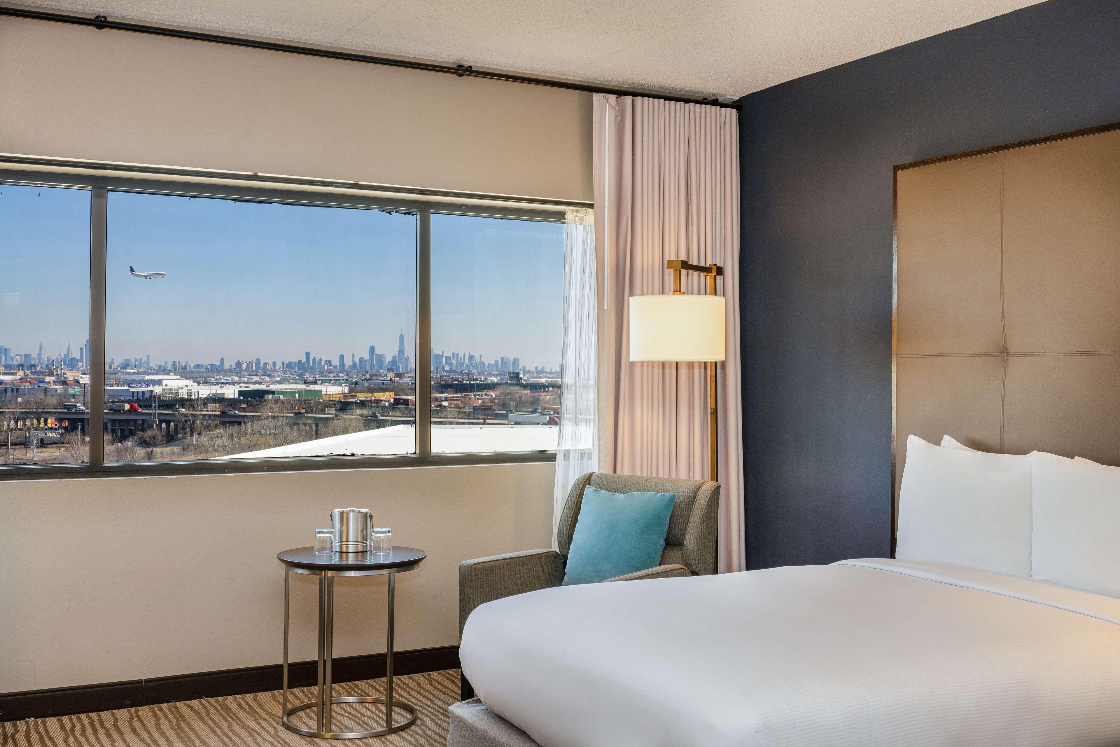 Une chambre d’hôtel avec une grande fenêtre, un lit, une chaise, une petite table et une lampe.