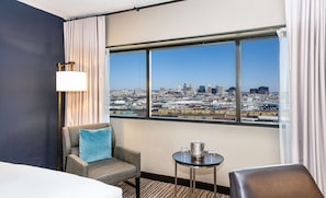 Habitación de hotel con un ventanal grande, una cama, una silla, una mesita con jarras y vasos, y una lámpara.