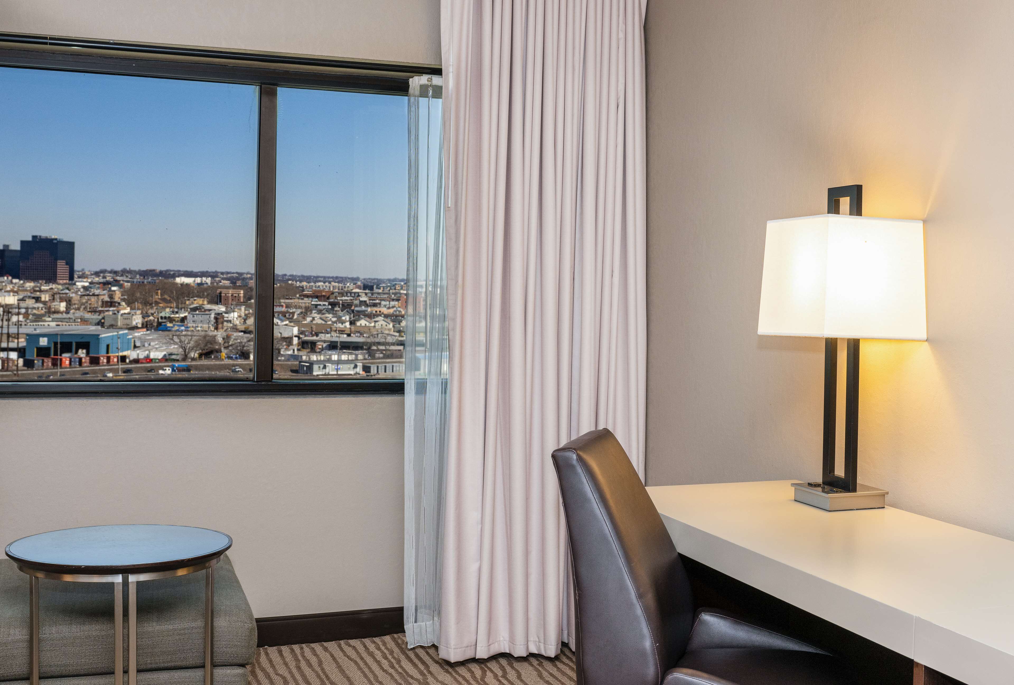 Une chambre d’hôtel avec vue sur la ville, une table ronde, une chaise, un bureau et une lampe.