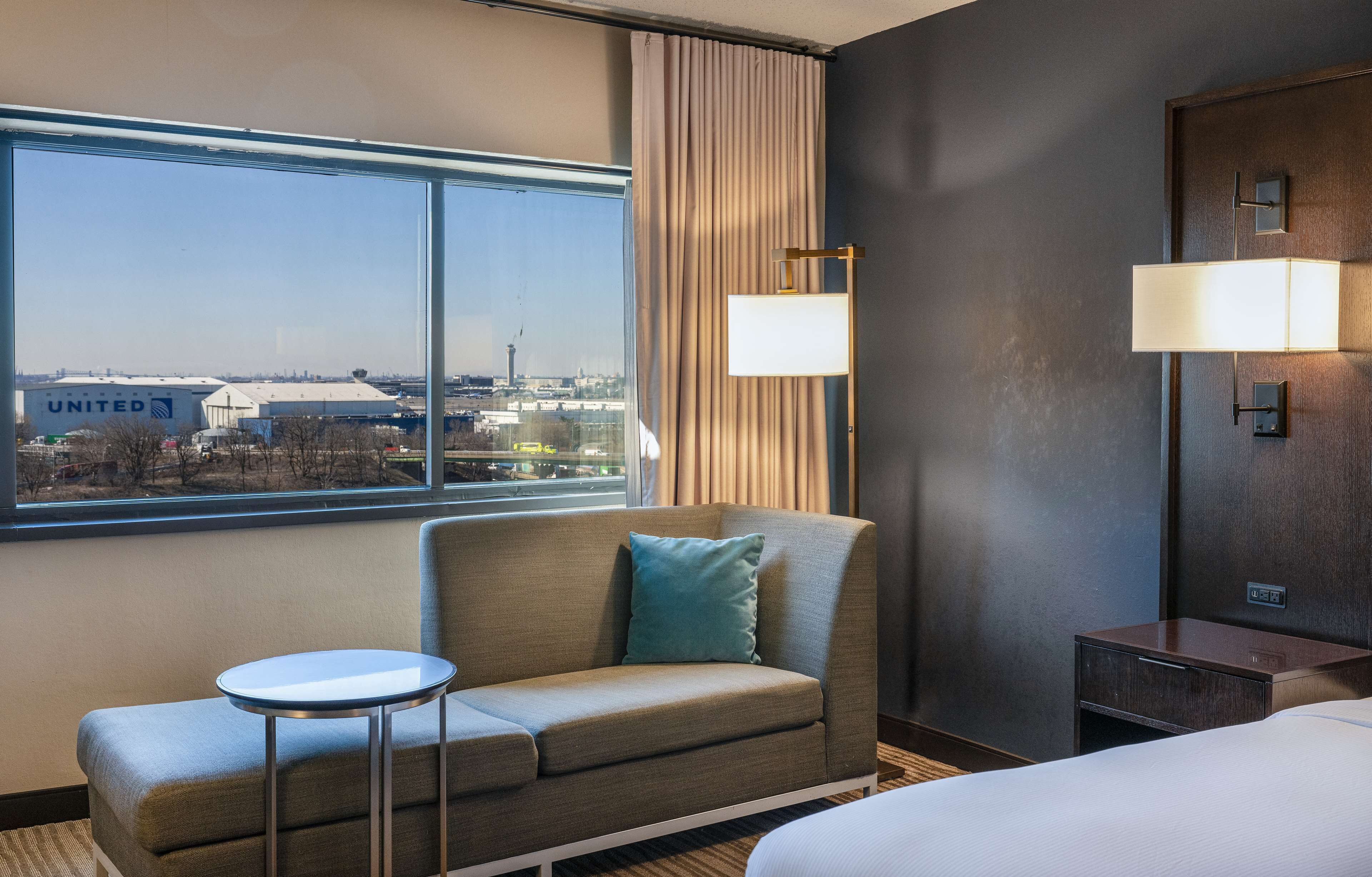 A hotel room with a large window, a sofa, a small table, a bed, and a nightstand with lamps.