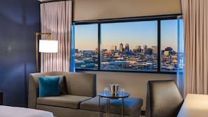 A hotel room with a large window offering a cityscape view, a sofa, a small table with a pitcher and glasses, and a lamp.
