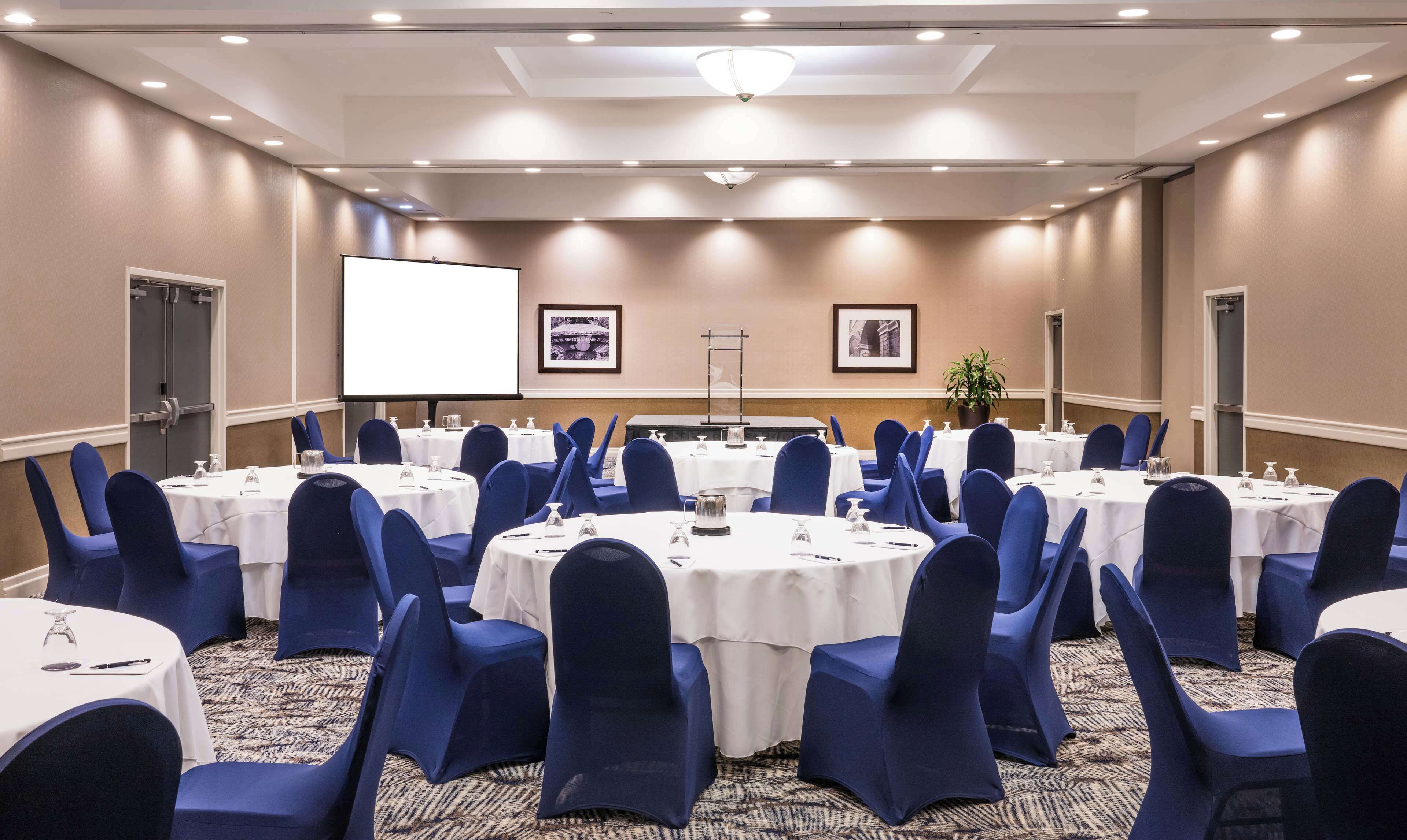 A conference room with round tables, blue chairs, white tablecloths, a projector screen, and framed pictures on the walls.