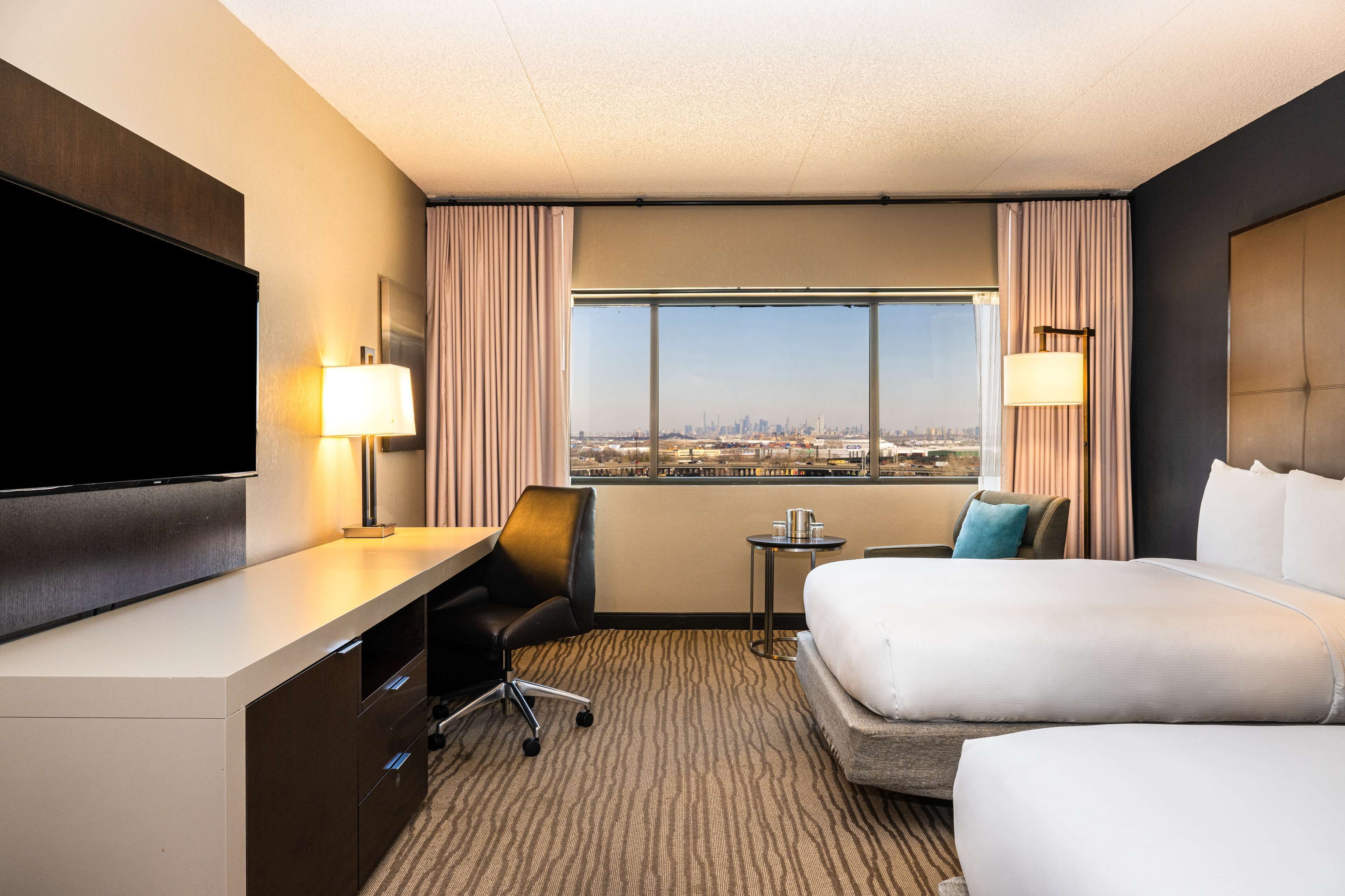 A hotel room with a large bed, a desk with a chair, a TV, a small table, and a view of the city.