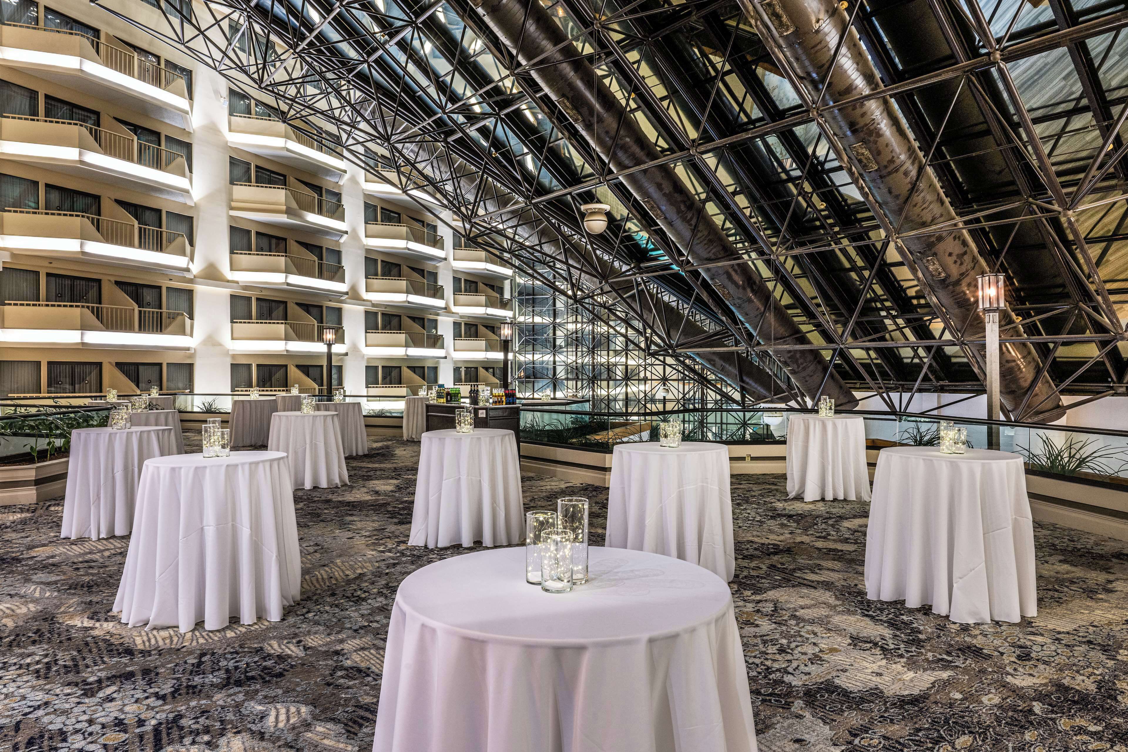 Un atrium d’hôtel avec de nombreuses tables rondes disposées pour les repas, un haut plafond avec une structure métallique et une vue sur les balcons extérieurs de l’immeuble.