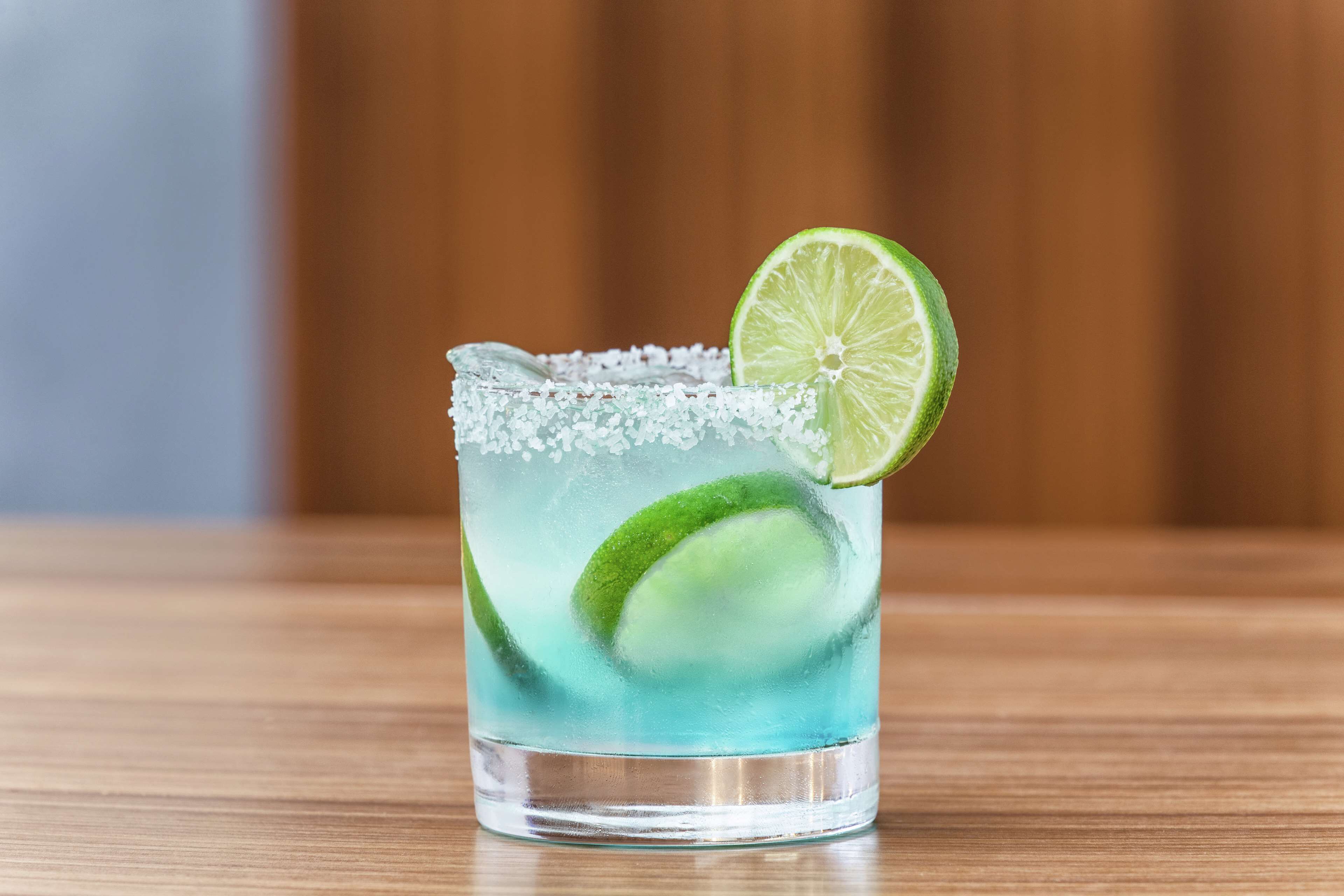A clear glass with a salted rim, filled with a blue liquid and lime slices, on a wooden surface.