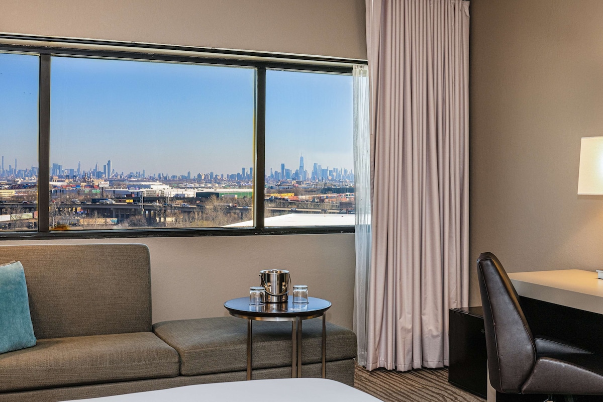 A hotel room with a large window offering a city view, a small table with a lamp, and a sofa.