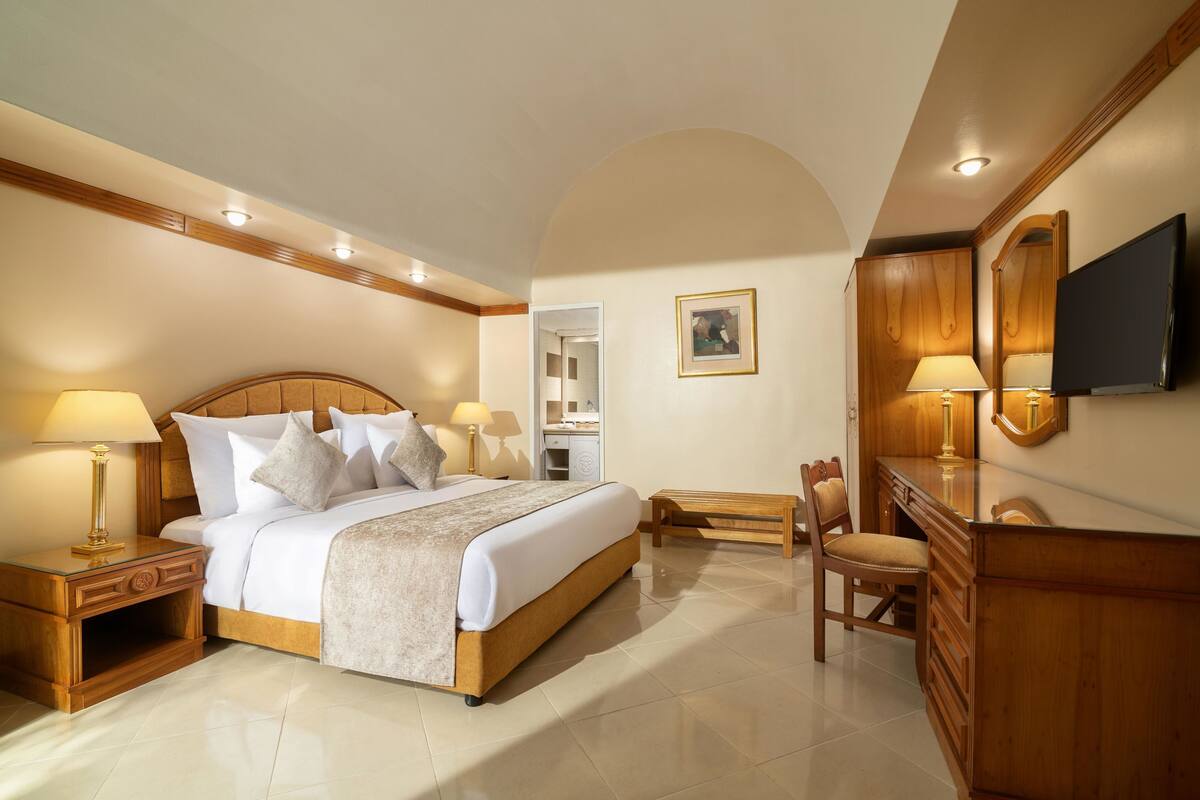 A hotel room with a large bed, bedside tables, a desk, a chair, a TV, and a mirror.