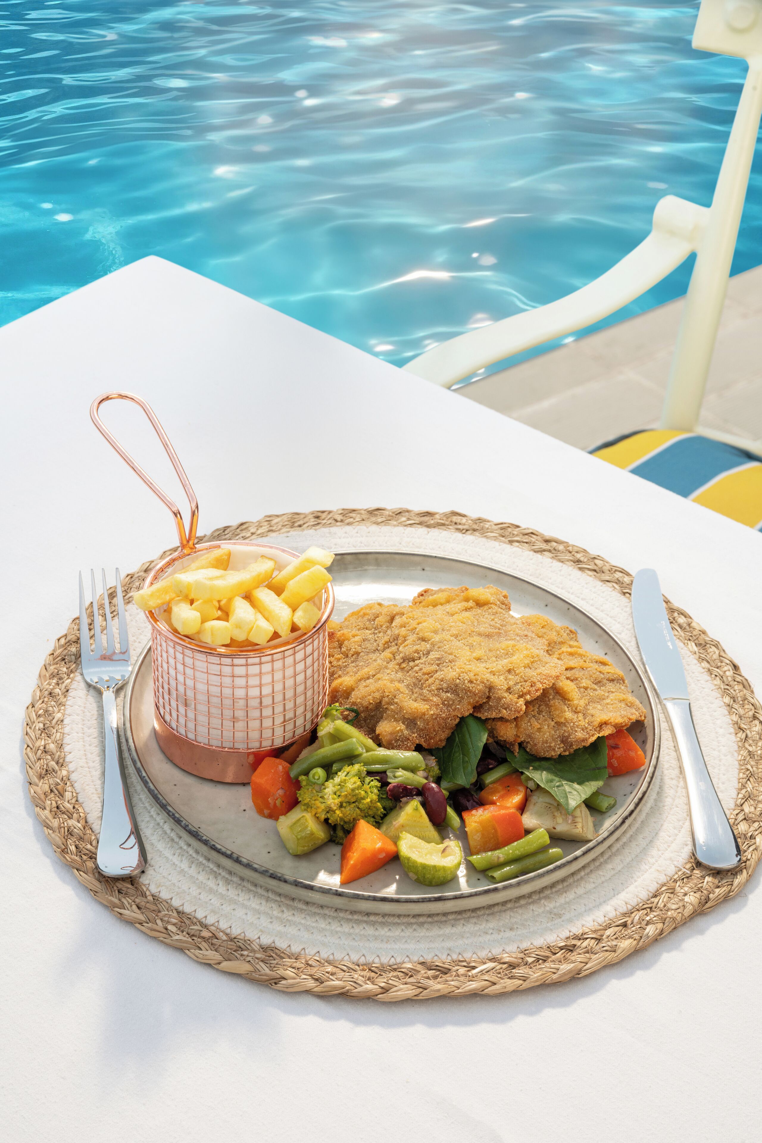 A meal of breaded fish, vegetables, and fries by a poolside table.