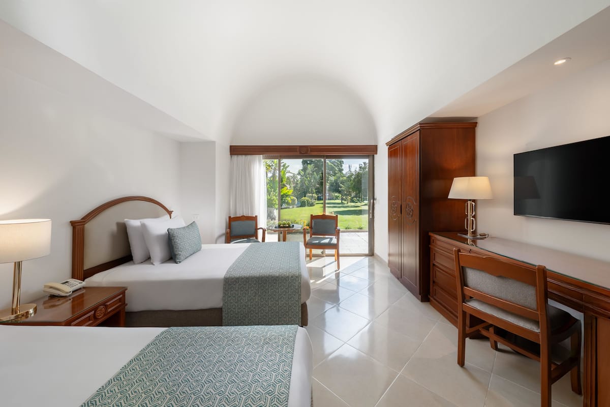 A hotel room with two beds, a desk, a TV, and a balcony with a view of greenery.