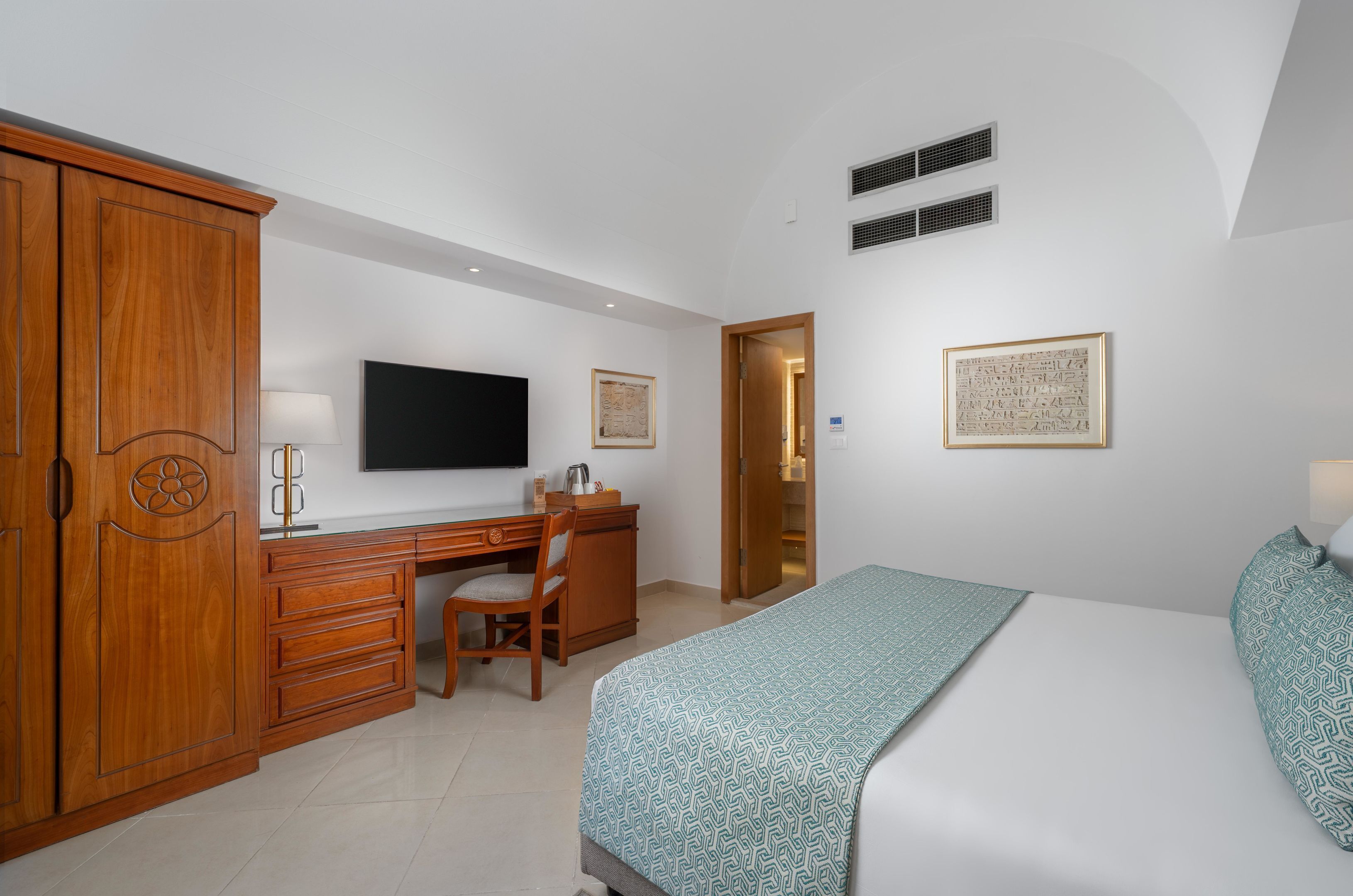 A hotel room with a wooden wardrobe, a desk with a chair, a bed with a patterned bedspread, a flat-screen TV, and two wall-mounted air vents.