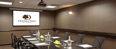 A conference room with a long table, chairs, a projector screen, and a logo on the wall.