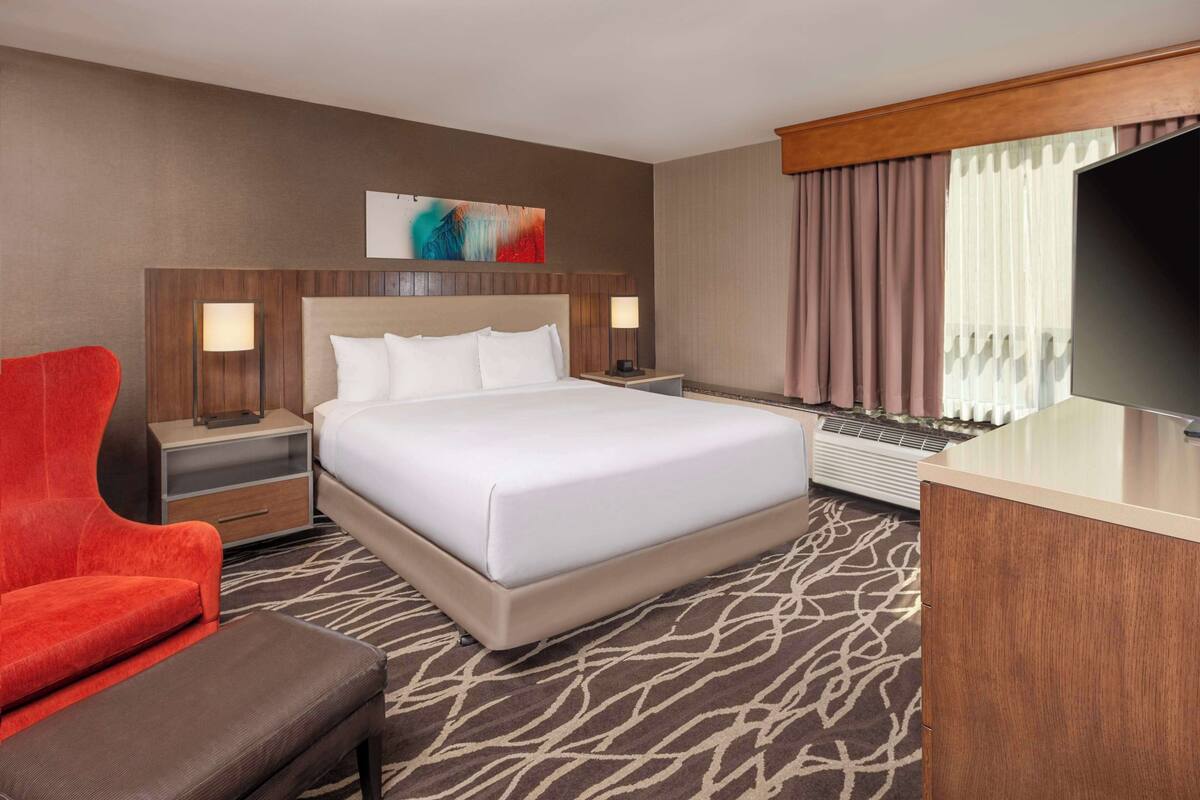 Une chambre d’hôtel avec un grand lit, un bureau, une télévision, un fauteuil rouge et une moquette à motifs.