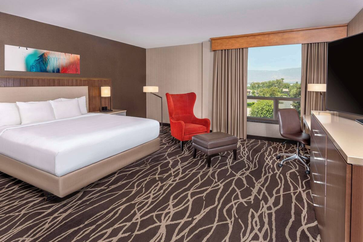 Une chambre d’hôtel avec un grand lit, un fauteuil rouge, un bureau avec une télévision et une vue sur l’extérieur.