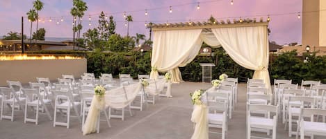 An outdoor wedding venue with white chairs arranged in rows, draped with white fabric and floral centerpieces.