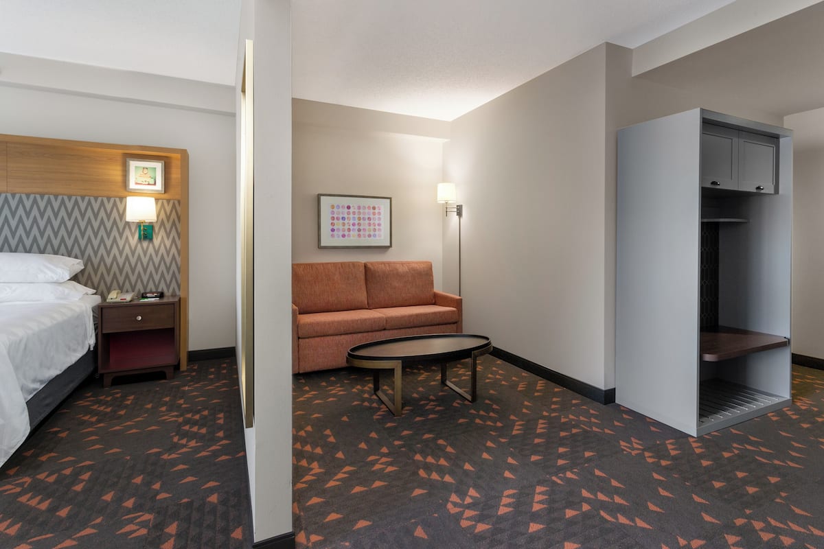 A hotel room with a bed, a sofa, a small table, a nightstand, a wall-mounted lamp, and a wardrobe.