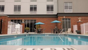 Ein Schwimmbecken mit einem Backsteingebäude im Hintergrund, Sitzgelegenheiten im Freien und Sonnenschirmen.