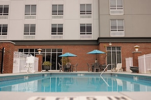 A swimming pool with a brick building in the background, outdoor seating, and umbrellas.