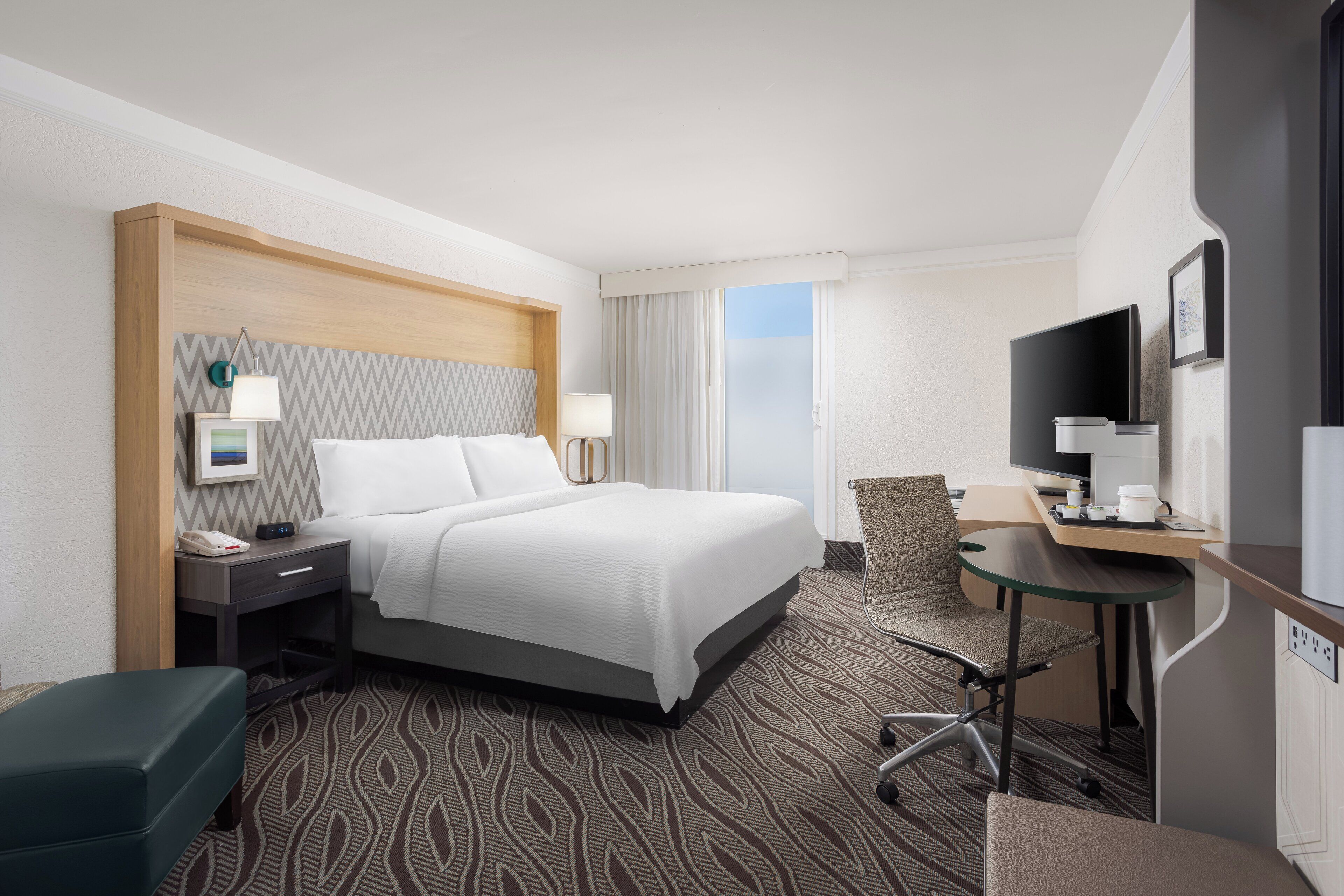 A hotel room with a bed, bedside table, lamp, chair, desk, TV, and a view of the outdoors.