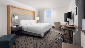 A hotel room with a bed, bedside table, lamp, chair, desk, TV, and a view of the outdoors.