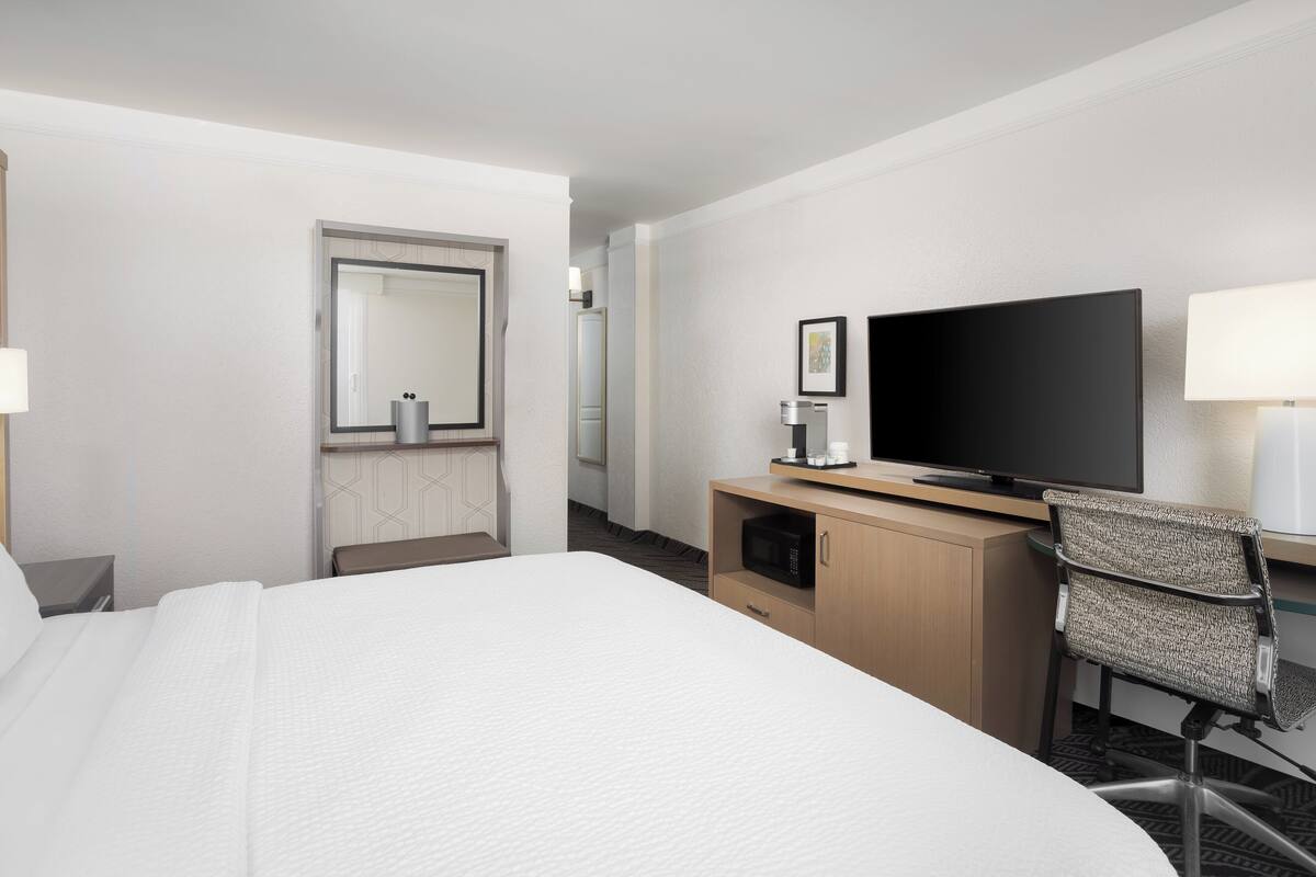 A hotel room with a large bed, a desk with a flat-screen TV, a chair, a microwave, and a door leading to another room.