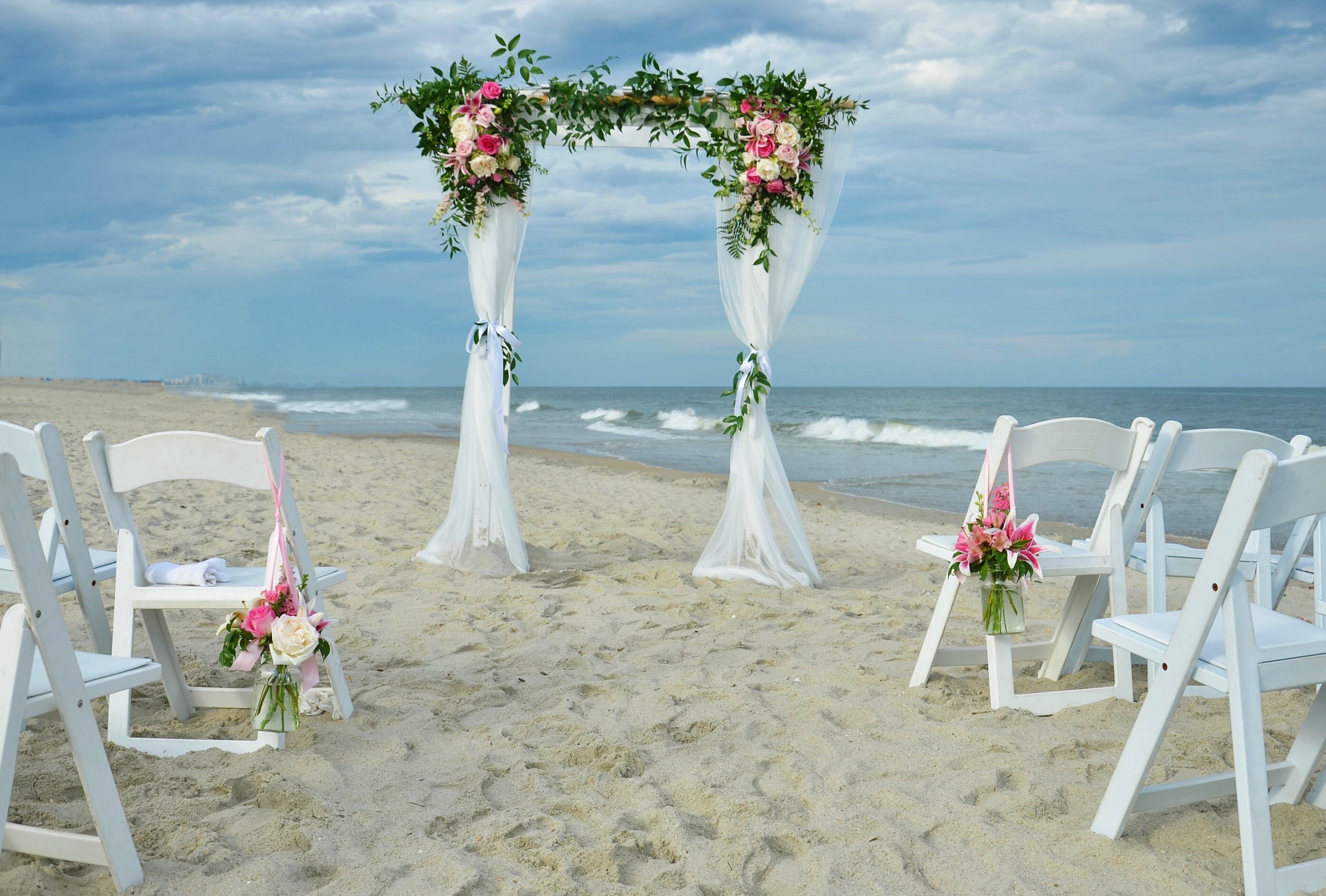 A beach wedding setup with white chairs and a floral arch.