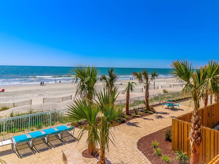 Vista de la playa con valla de madera, palmeras y un camino pavimentado con bancos.