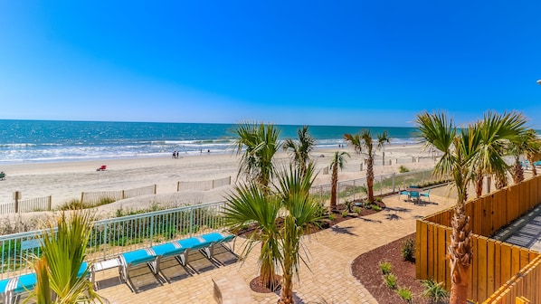 A beach view with a wooden fence, palm trees, and a paved walkway with benches.