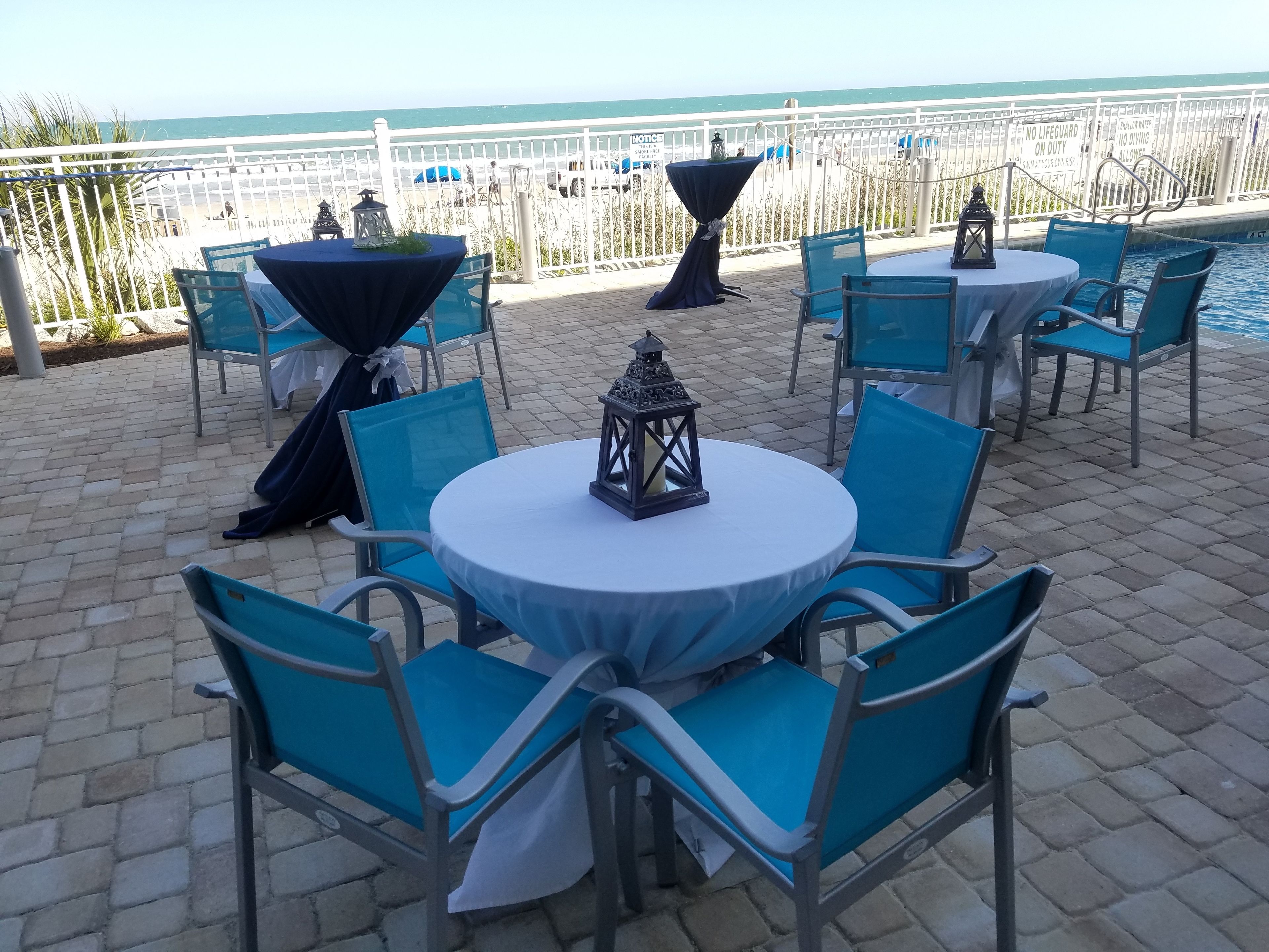 A seaside patio with round tables and blue chairs, overlooking the ocean.