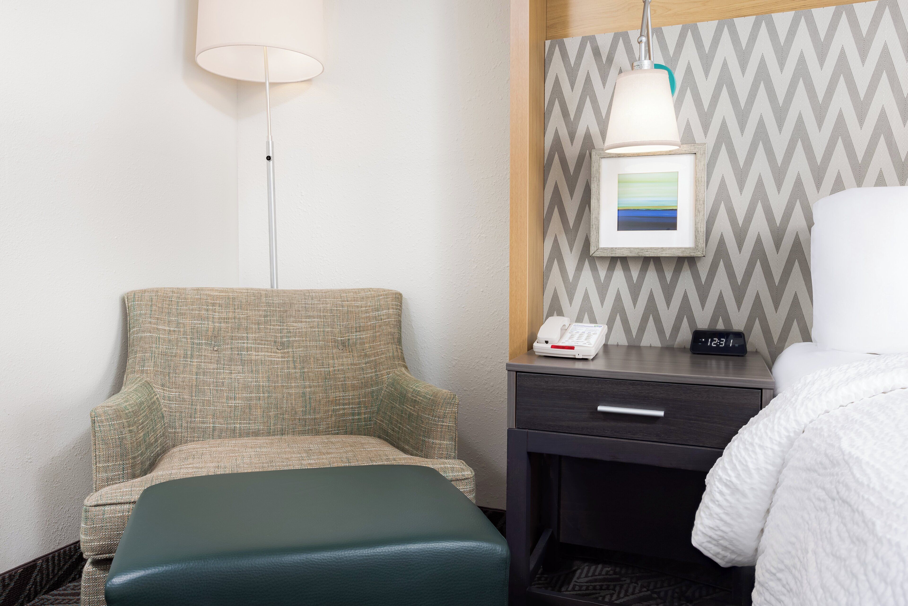 A hotel room with a green upholstered chair, a bedside table with a lamp, a framed picture, and a digital clock displaying 12:31.