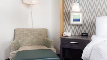 A hotel room with a green upholstered chair, a bedside table with a lamp, a framed picture, and a digital clock displaying 12:31.
