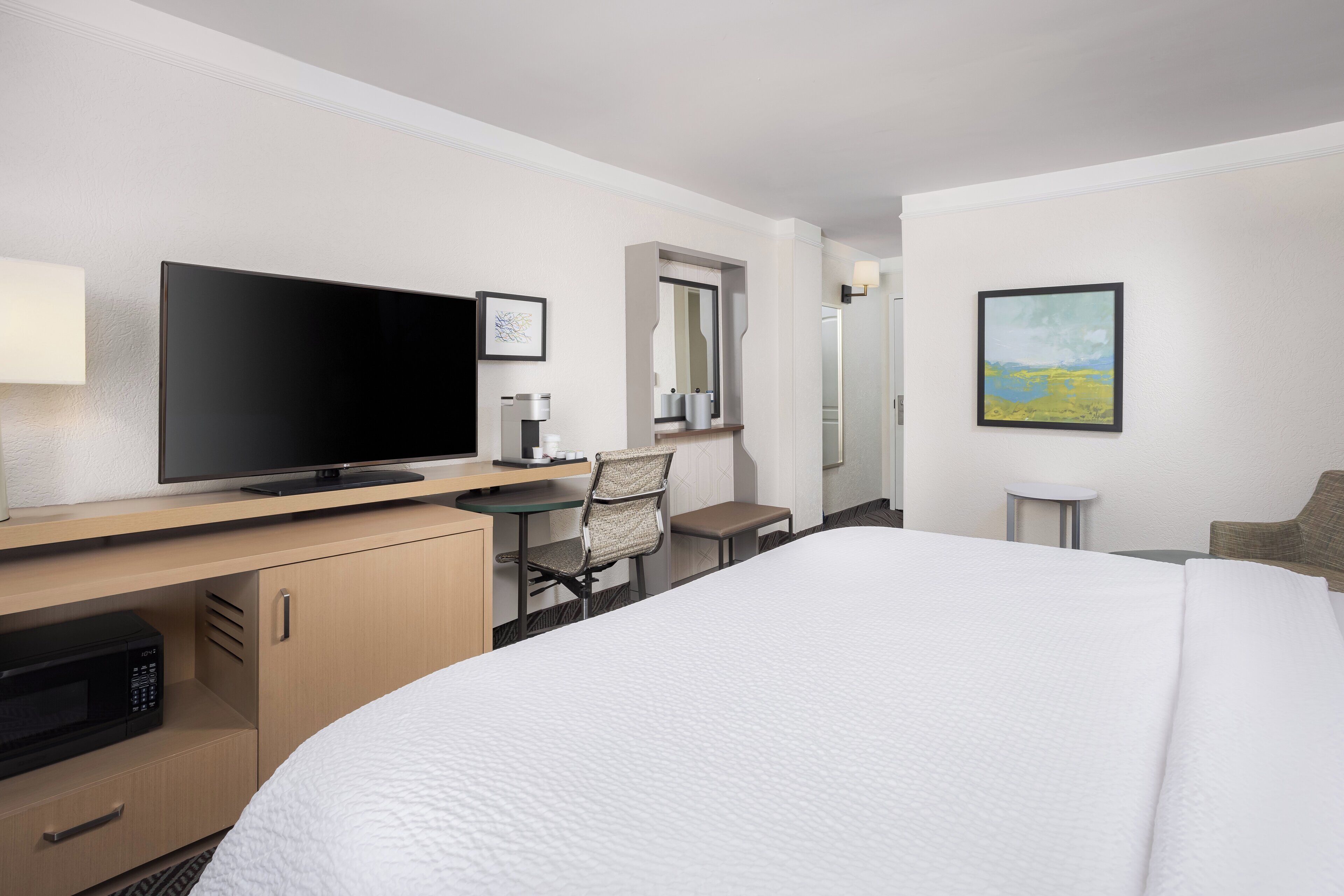 A hotel room with a bed, a desk with a flat-screen TV, a chair, a small table, and a painting on the wall.