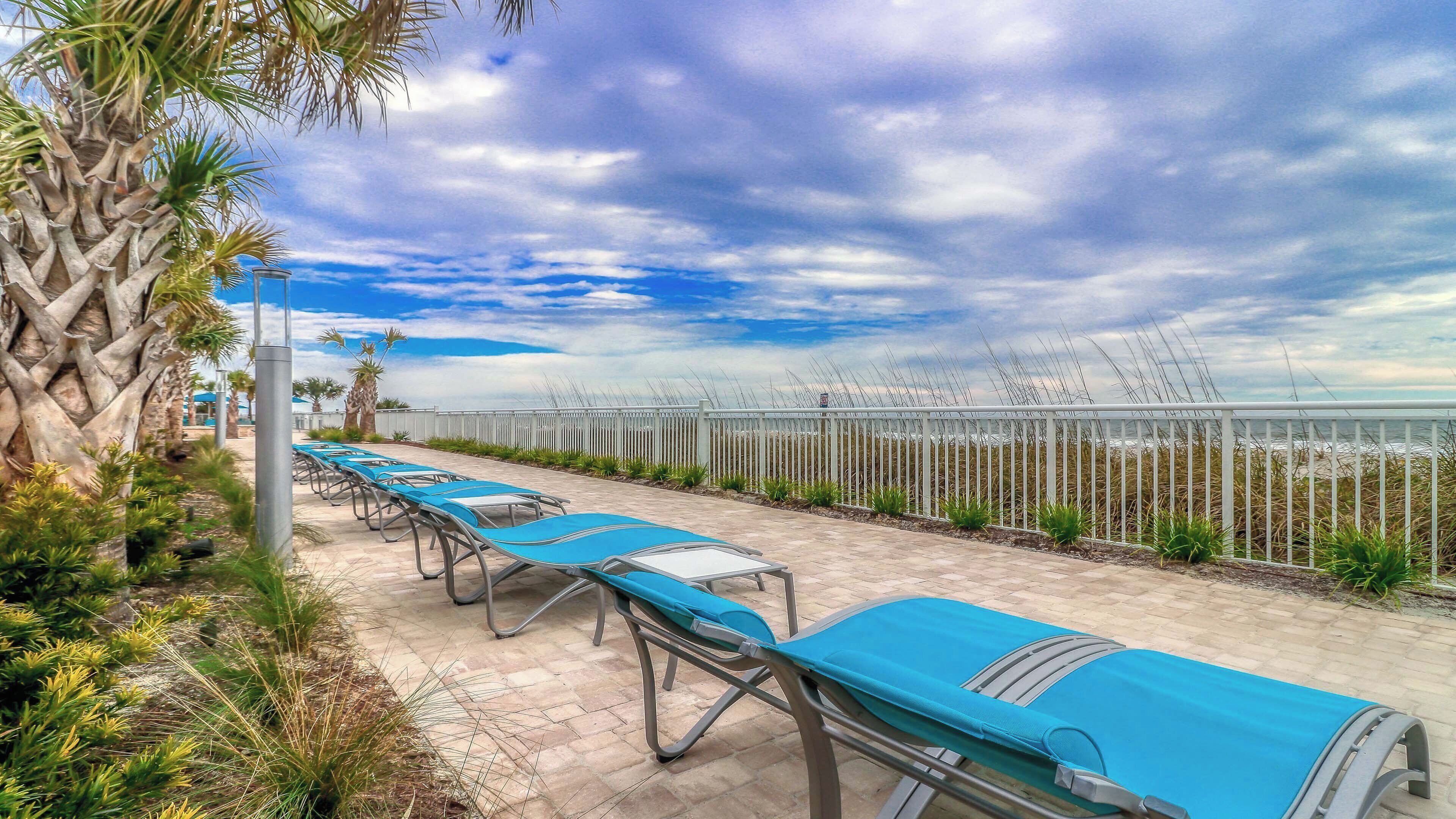A row of blue lounge chairs aligned along a paved walkway, with a white fence and palm trees in the background.