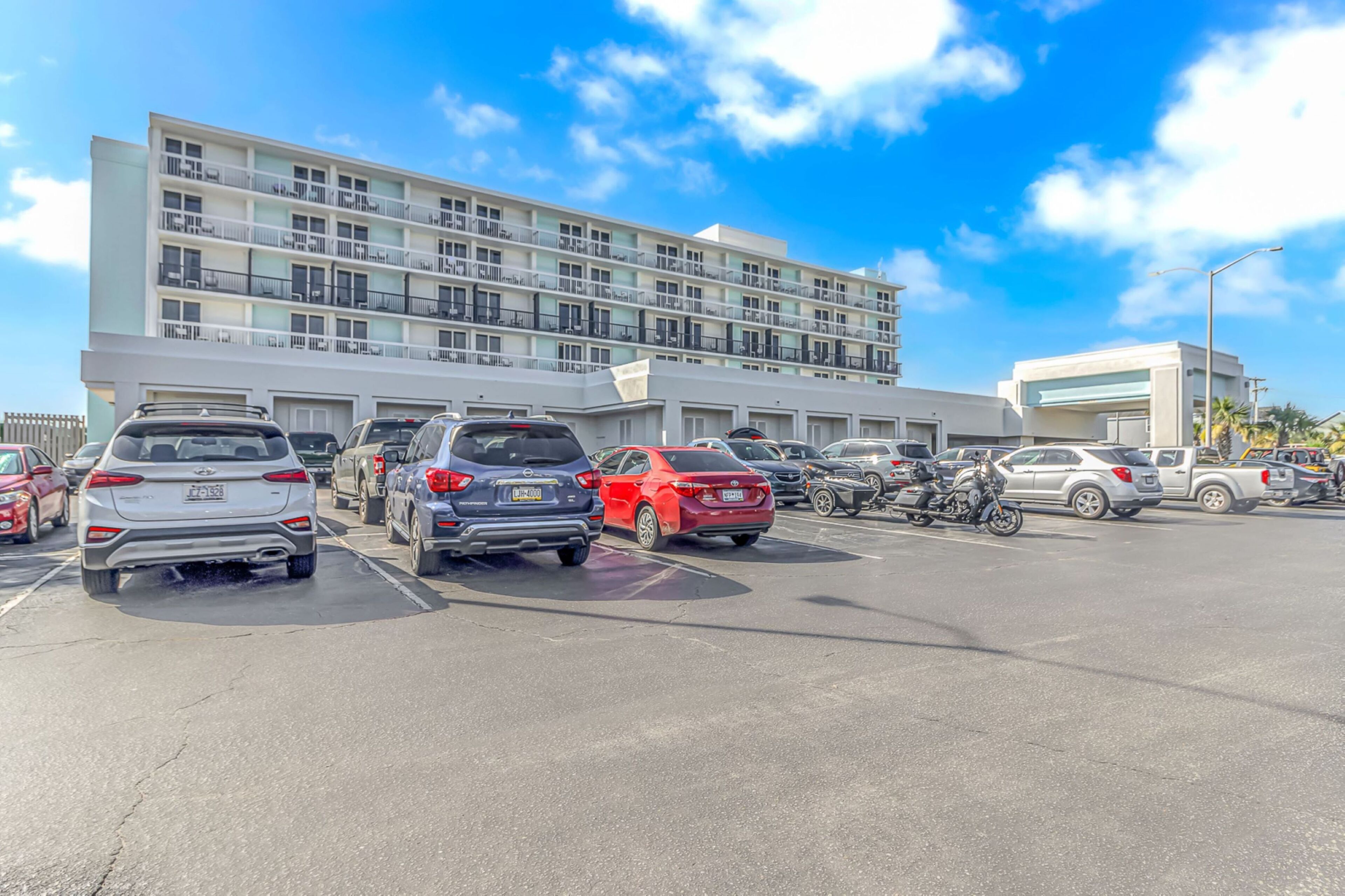 A parking lot with multiple cars and a motorcycle, a multi-story building with balconies, and a clear blue sky.