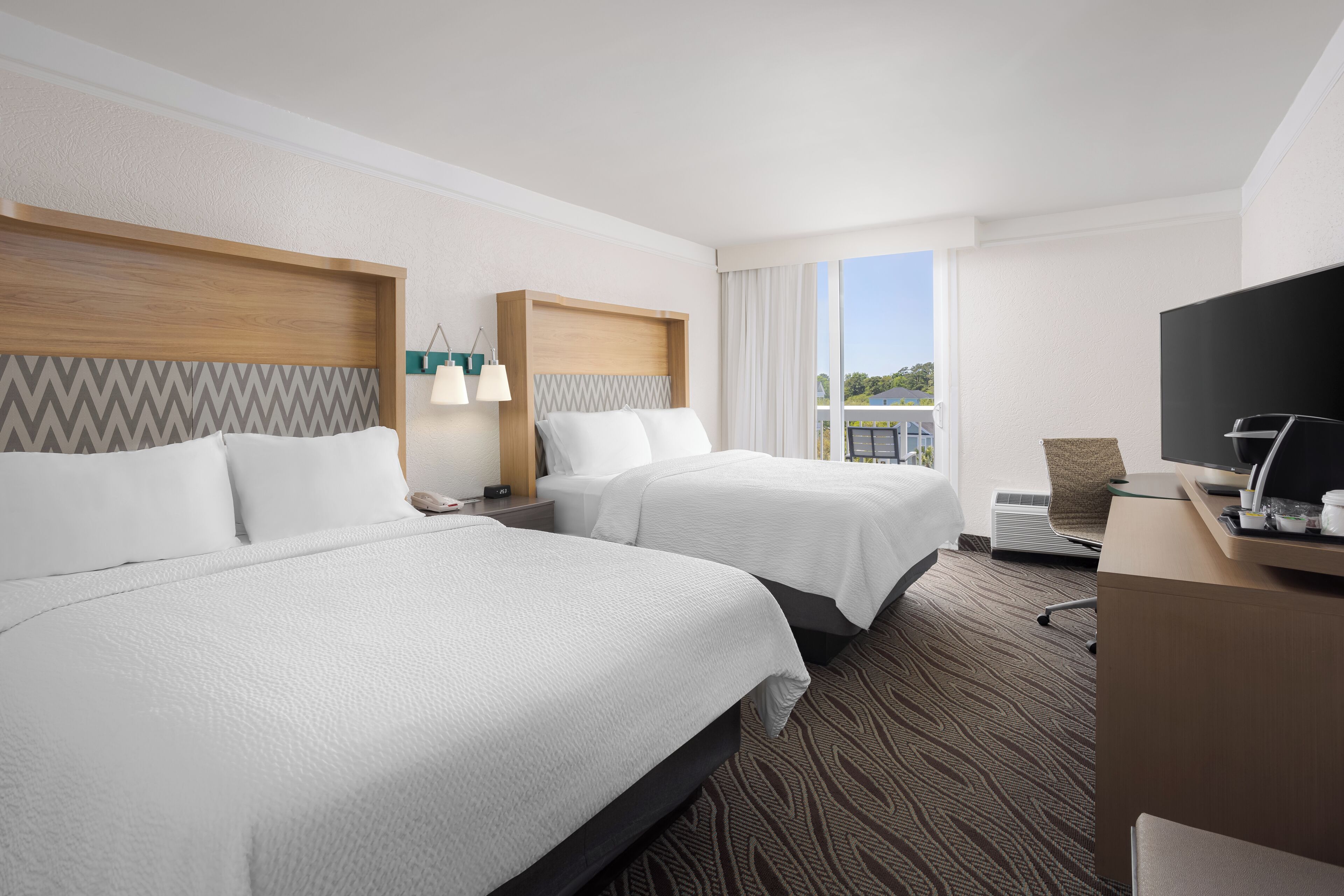 A hotel room with two beds, a desk with a television, and a window with a view of buildings.