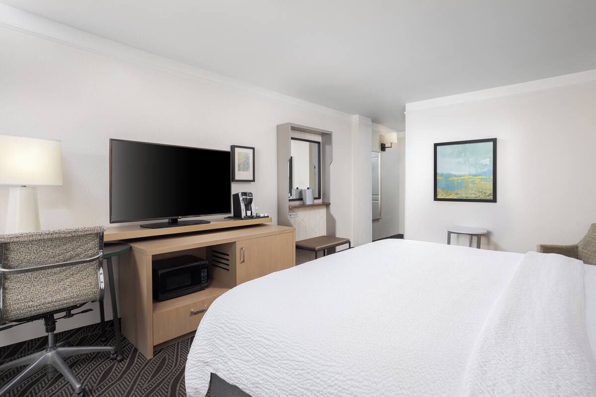 A hotel room with a large bed, a flat-screen TV, a desk with a lamp, a chair, a microwave, and a small table.