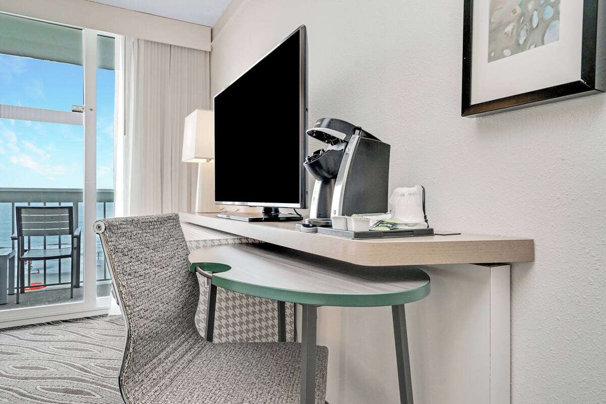 A hotel room with a desk, a chair, a flat-screen TV, a lamp, and a framed picture on the wall.