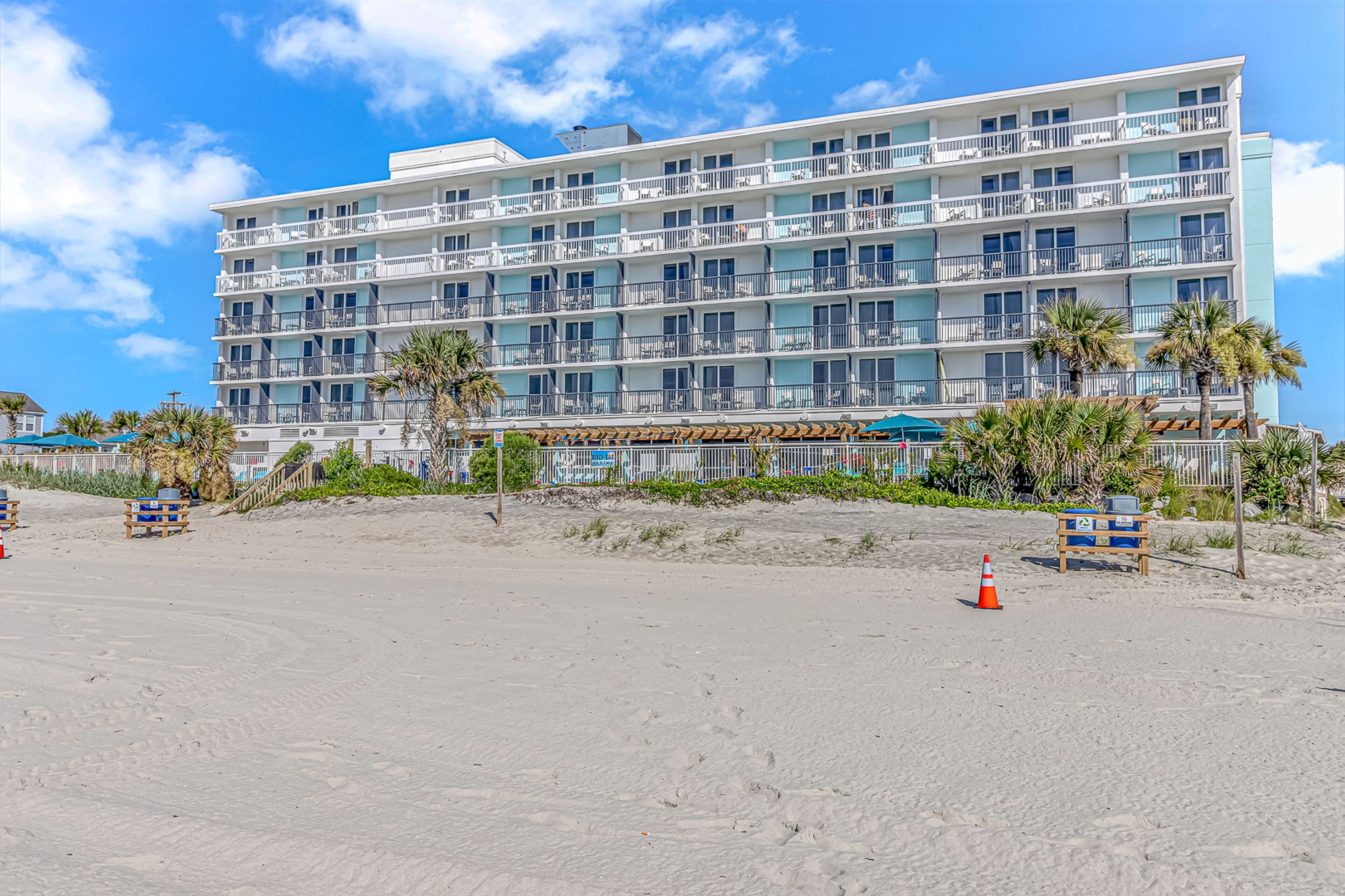 A multi-story hotel with balconies, located on a sandy beach with palm trees and beach chairs.