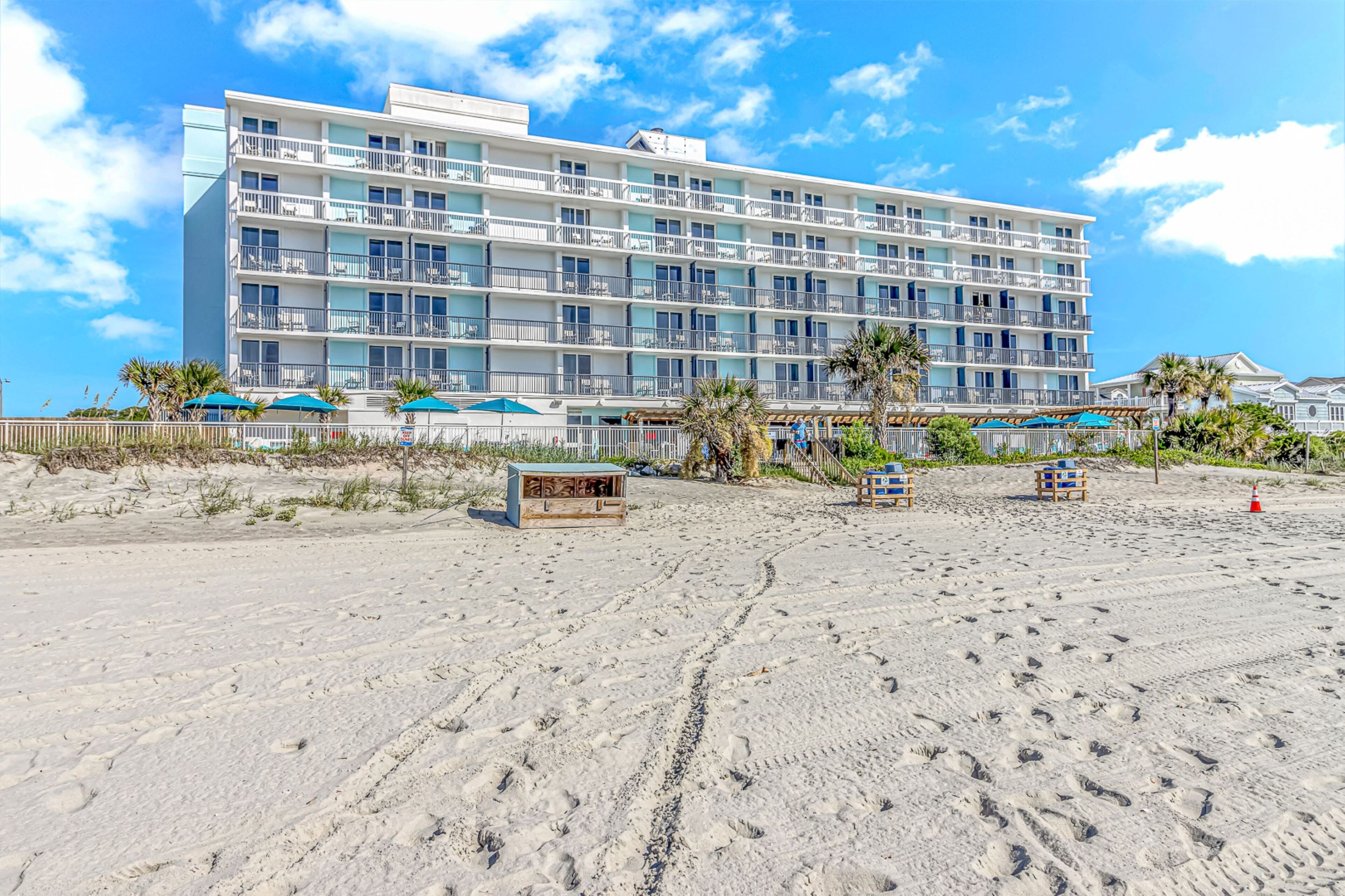 Un hotel frente a la playa con varios balcones, una playa arenosa y palmeras.