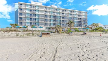 Un hotel frente a la playa con varios balcones, una playa arenosa y palmeras.