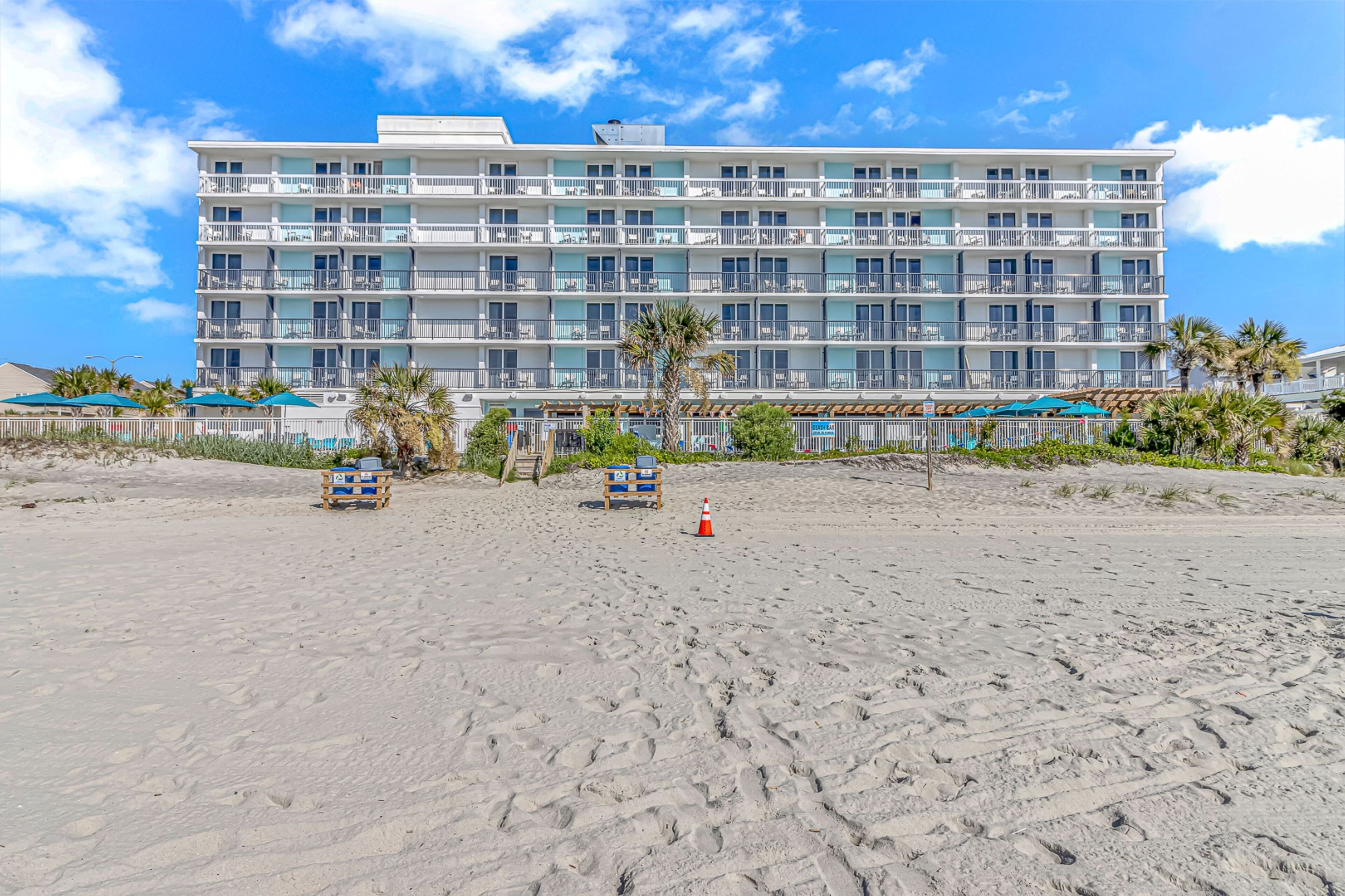 A multi-story hotel with balconies, located on a sandy beach with palm trees and beach chairs.