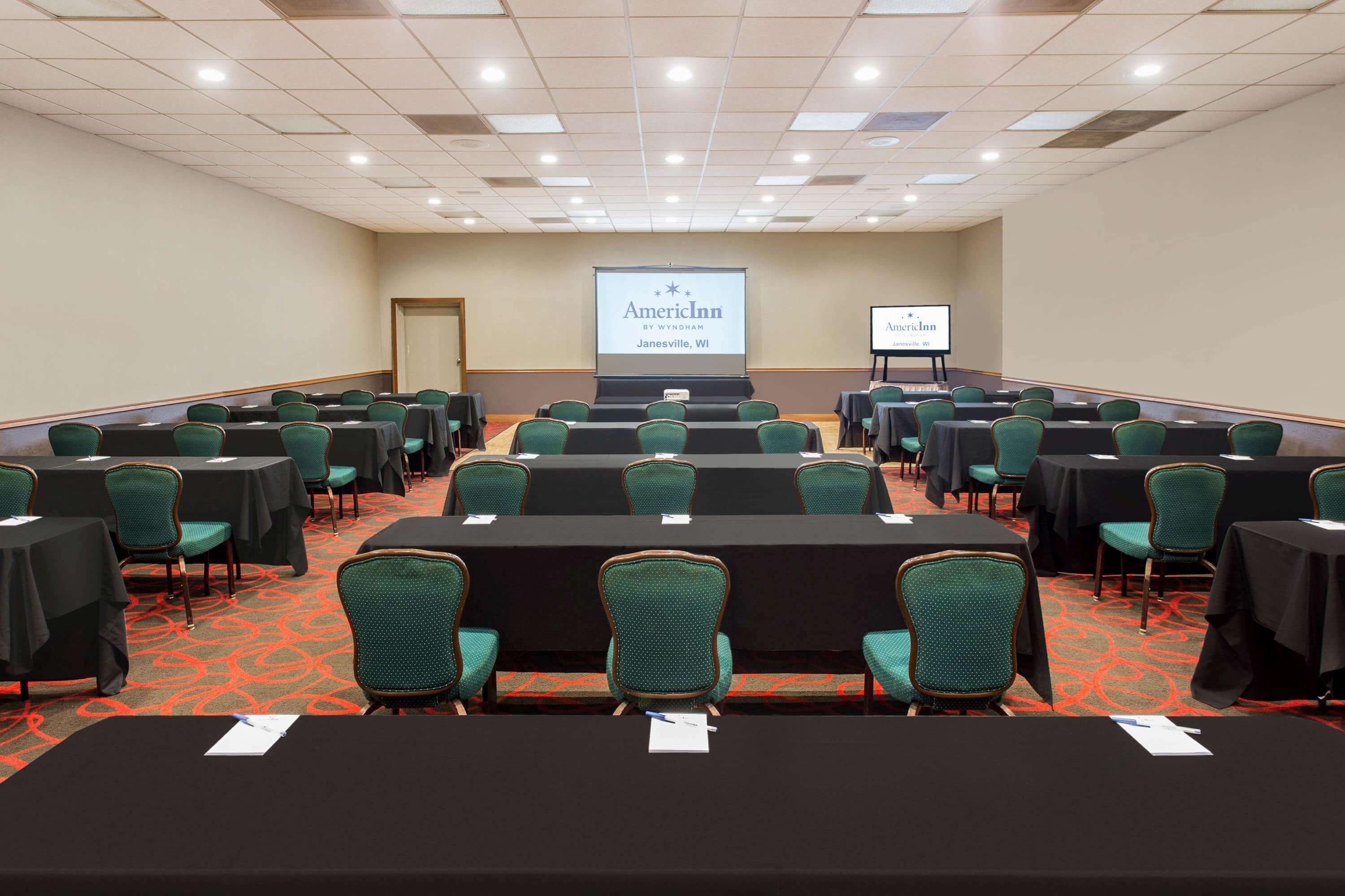 A conference room with rows of tables and chairs, a projector screen displaying 'American Inn by Wyndham Janesville, WI', and a carpeted floor.
