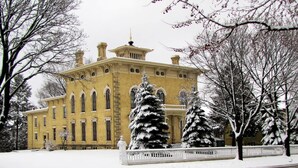 A historic yellow building with arched windows and a central tower, surrounded by snow-covered trees and a white fence.